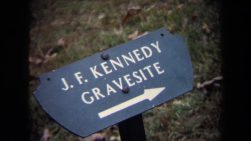 Close handheld 8mm frame of the green J.F. Kennedy Gravesite sign, tilted across grass with the white arrow visible.