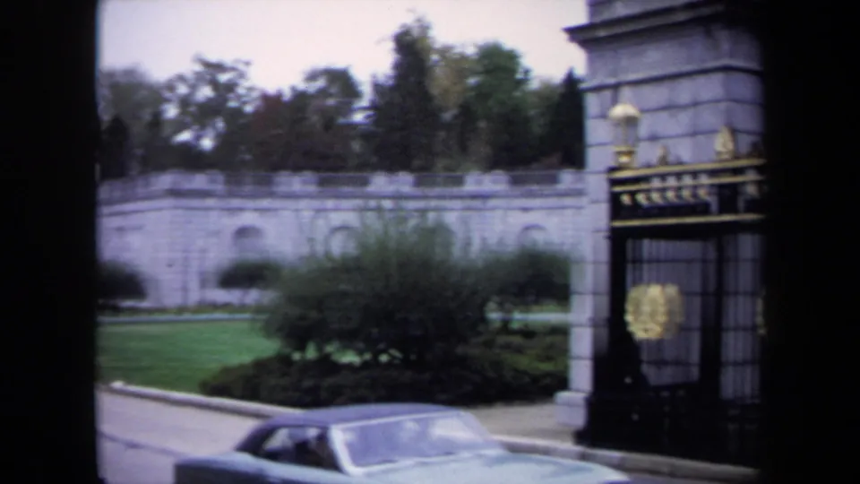 1970 Washington-area 8mm frame showing a stone entrance, black iron gate with gold details, shrubs, lawn, and a parked car.