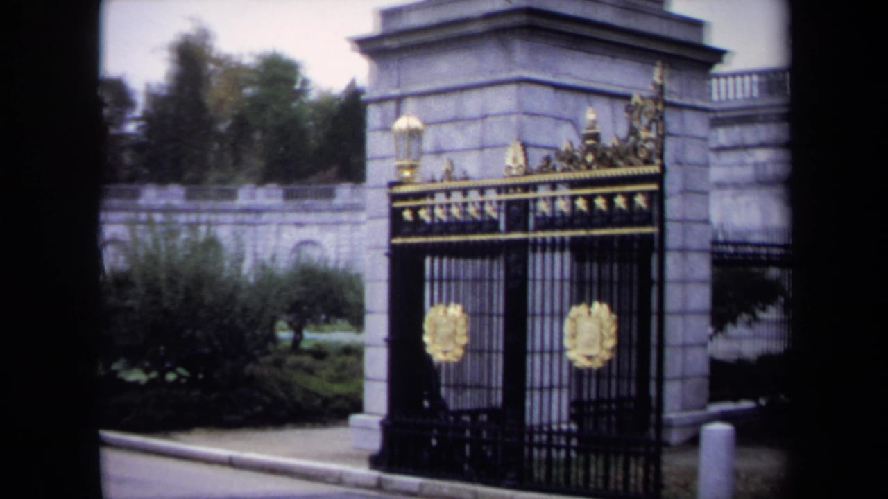 1970 Washington, DC home-movie frame showing a formal stone gate, black iron fencing, and gold emblem near landscaped grounds.