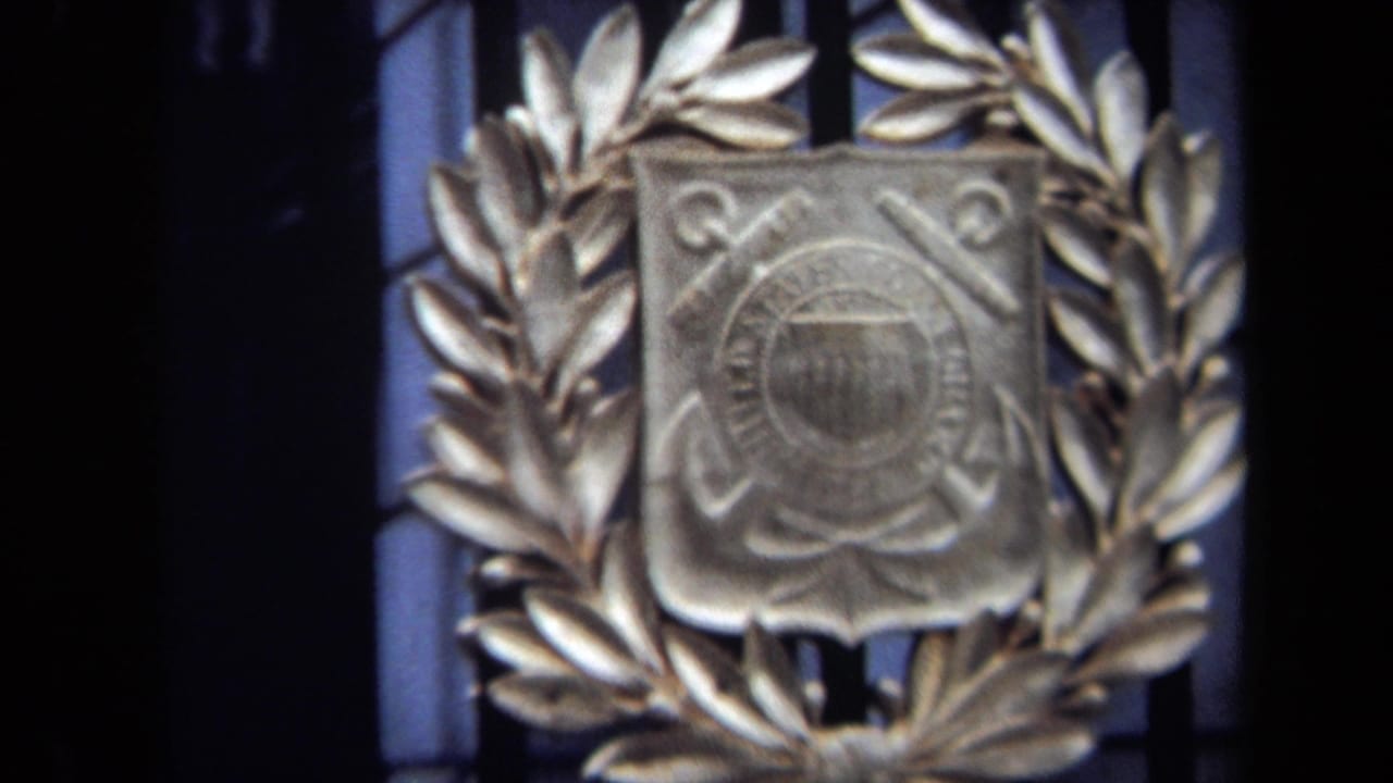 Close 1970 Washington, DC frame of a Coast Guard crest with laurel leaves, soft focus, and a dark barred background.