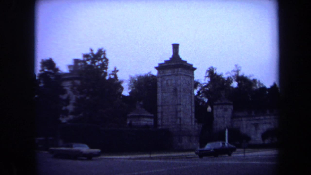 1970 Washington, DC 8mm frame showing a formal stone gateway with pillars, trees, traffic lights, and passing sedans.