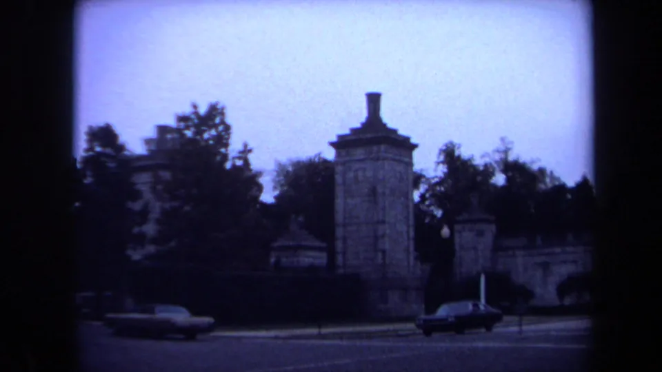 1970 Washington, DC 8mm frame showing a formal stone gateway with pillars, trees, traffic lights, and passing sedans.