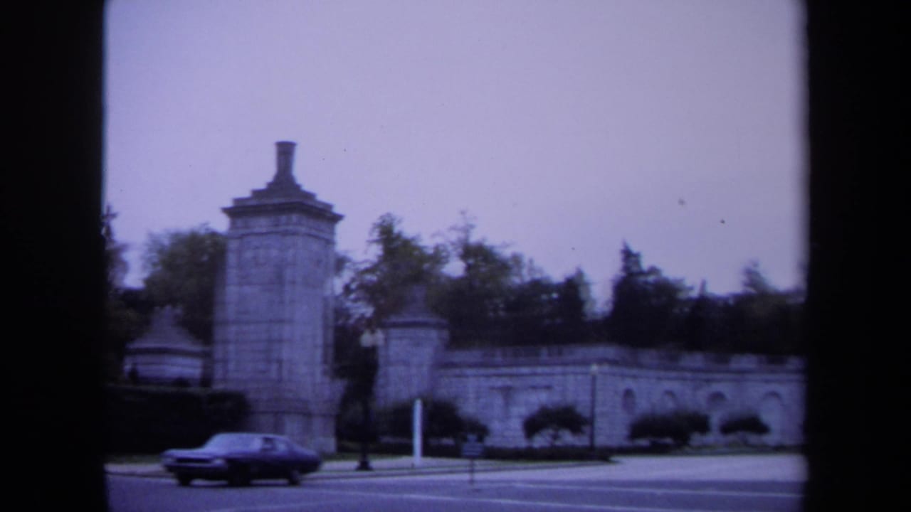 1970 Washington, DC view of a sedan passing a stone gate pillar, arched wall, hedges, and traffic signal.