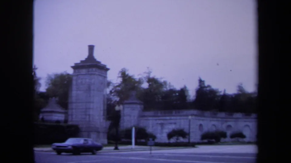 1970 Washington, DC view of a sedan passing a stone gate pillar, arched wall, hedges, and traffic signal.
