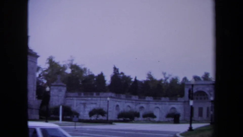 1970 Washington, DC 8mm frame with stone arched wall, roadside hedges, traffic signal, and partial sedan in the foreground.
