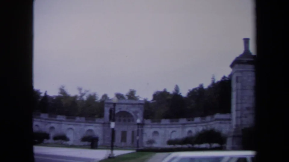 1970 Washington, DC frame showing a stone gateway wall, trees, traffic lights, roadway, and the top of a passing car.