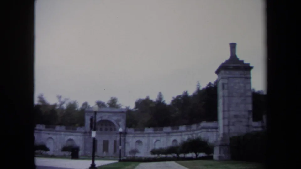 1970 Washington, DC home-movie frame showing a stone gateway, arched openings, trees, and a traffic light through dark borders.