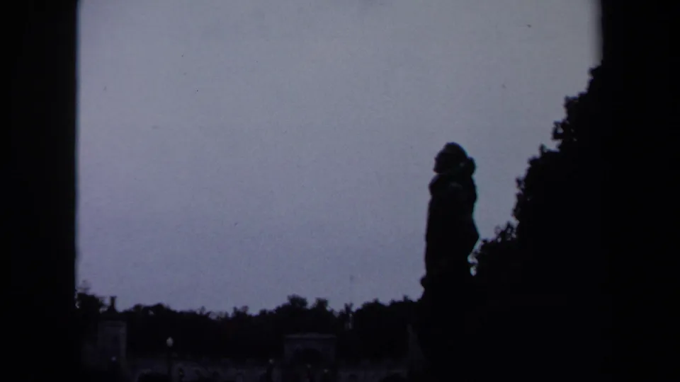 1970 Washington, DC home-movie frame showing a dark standing silhouette near foliage and distant stone architecture.