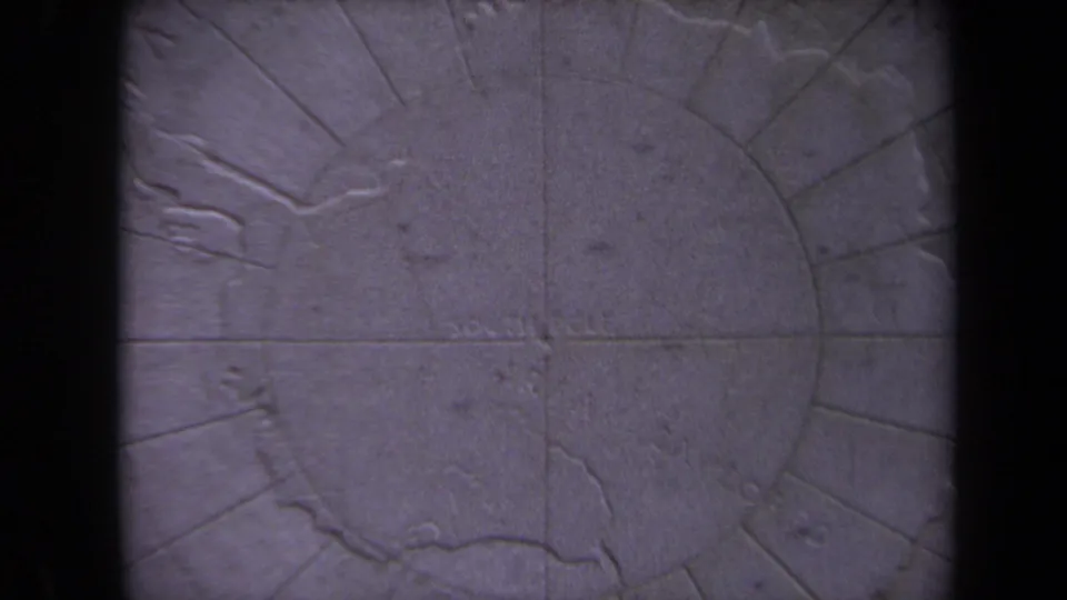 1970 Washington, DC home-movie frame showing a carved stone polar map surface with faint inscriptions and dark foreground bars.