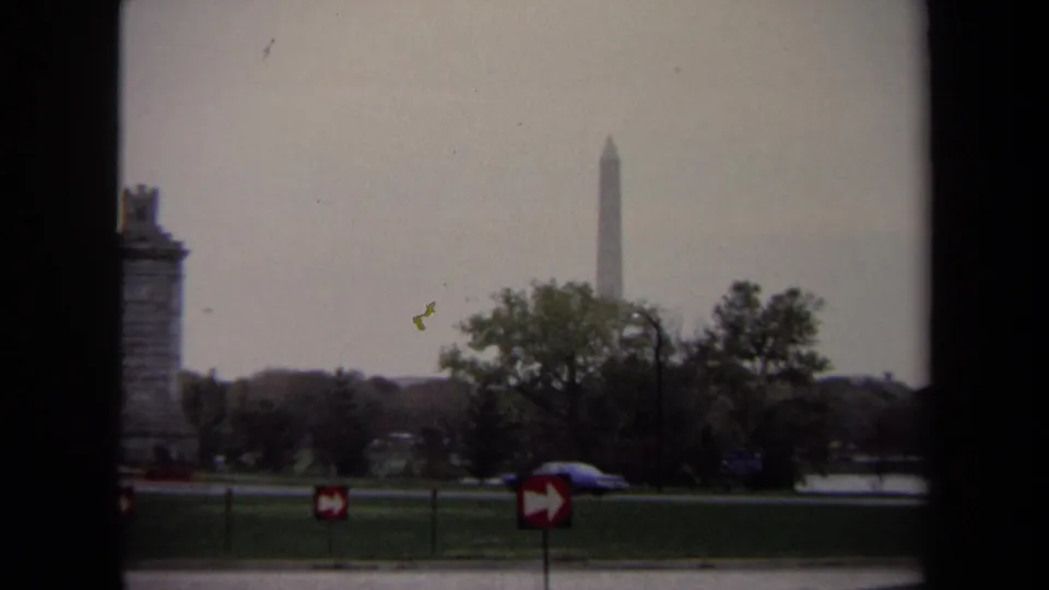 1970 Washington, DC home-movie frame with the Washington Monument rising in the distance beyond trees, road signs, grass, and passing cars.