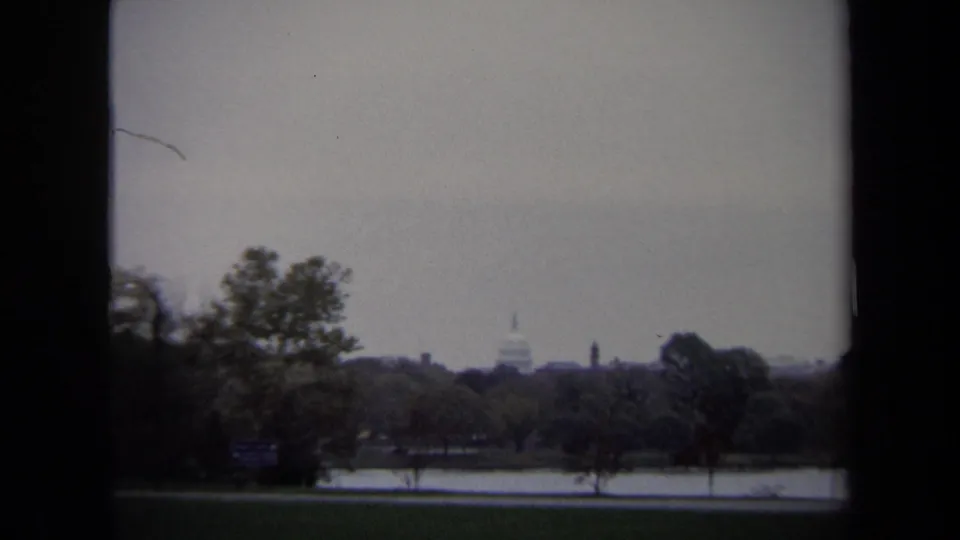 1970 Washington, DC view of the U.S. Capitol dome across a reflective body of water, with trees, parkland, and a hazy skyline.