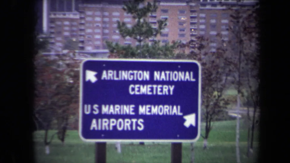 1970 Washington DC area roadside sign pointing toward Arlington National Cemetery, the U.S. Marine Memorial, and airports, with buildings behind.