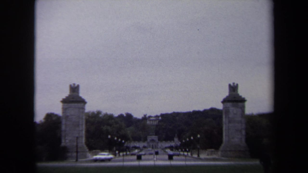 1970 Washington DC 8mm frame showing paired stone pylons beside a tree-lined road with cars and distant memorial architecture.