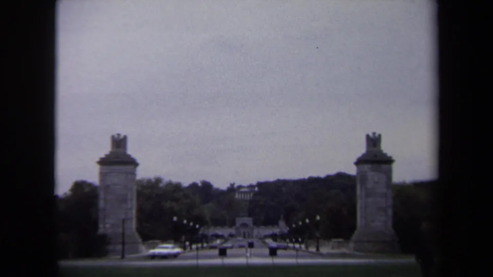 1970 Washington DC 8mm frame showing paired stone pylons beside a tree-lined road with cars and distant memorial architecture.