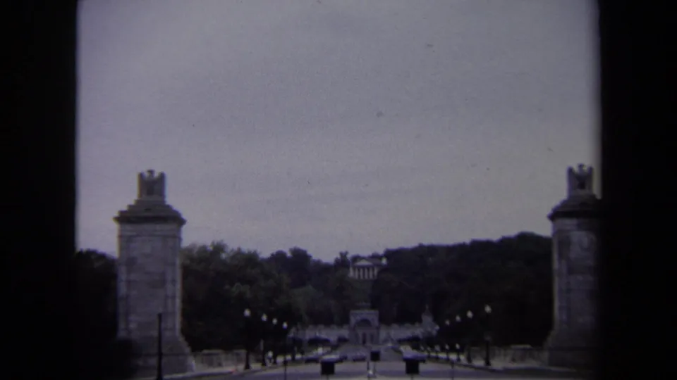 1970 Washington DC visitor-shot frame of stone pylons, a formal road approach, small pedestrians, cars, and a distant monument-like structure.