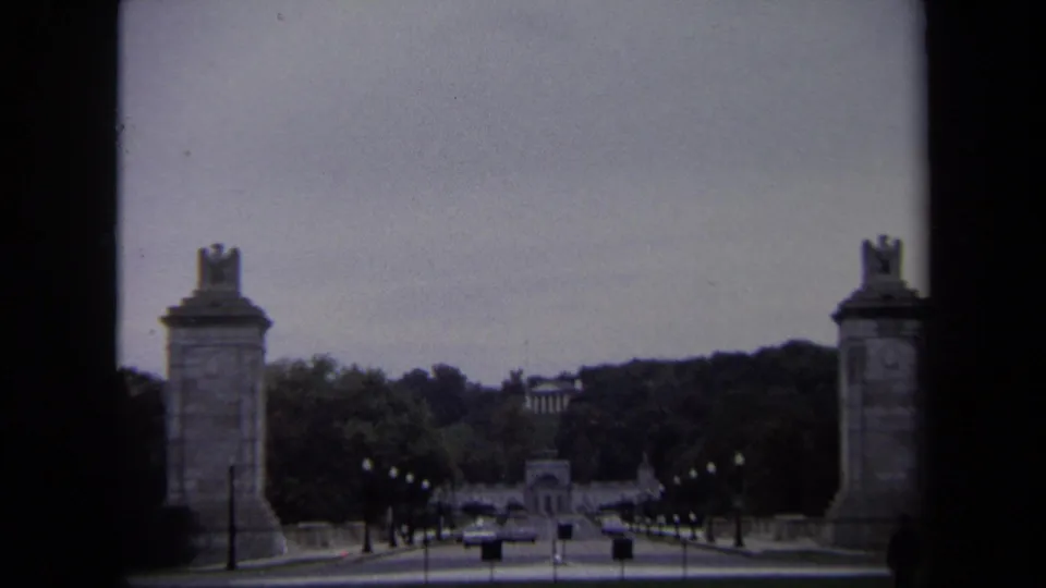 1970 Washington, DC 8mm frame showing eagle-topped stone pylons beside a road leading toward a distant neoclassical building.