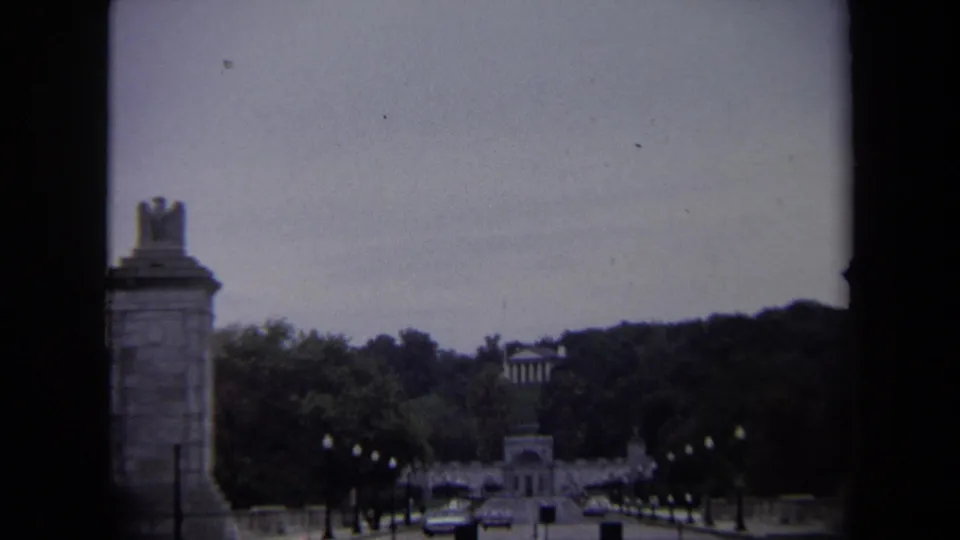 1970 Washington, DC home-movie view of cars on a tree-lined approach road between monumental stone pylons.
