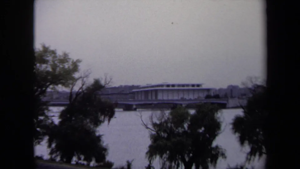1970 Washington, DC home-movie frame showing a river, shoreline trees, and a modern building on the far bank.