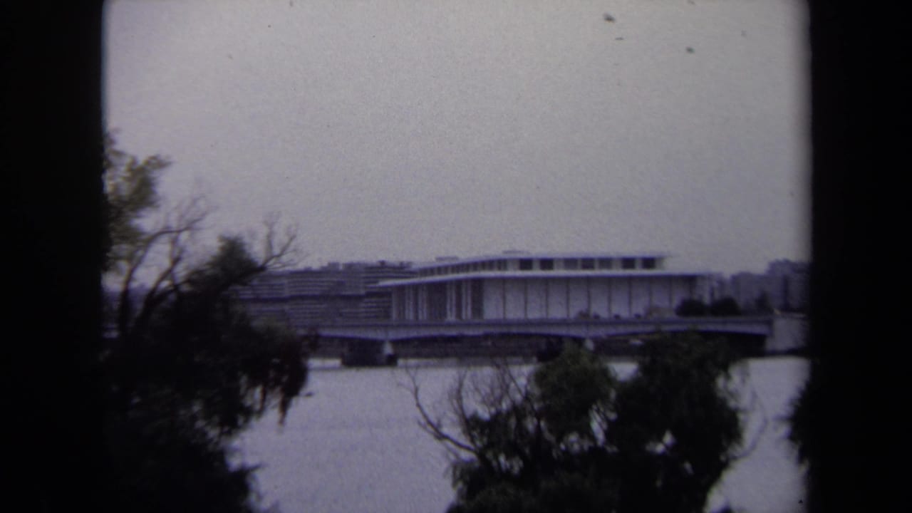 1970 Washington, DC 8mm frame showing a distant modernist riverfront structure and bridge or overpass through foreground trees.