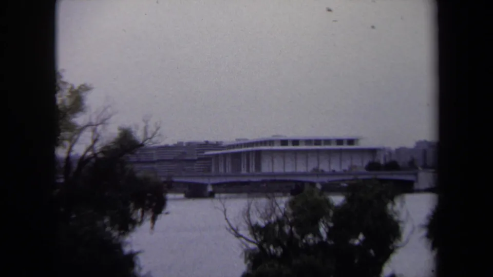 1970 Washington, DC 8mm frame showing a distant modernist riverfront structure and bridge or overpass through foreground trees.