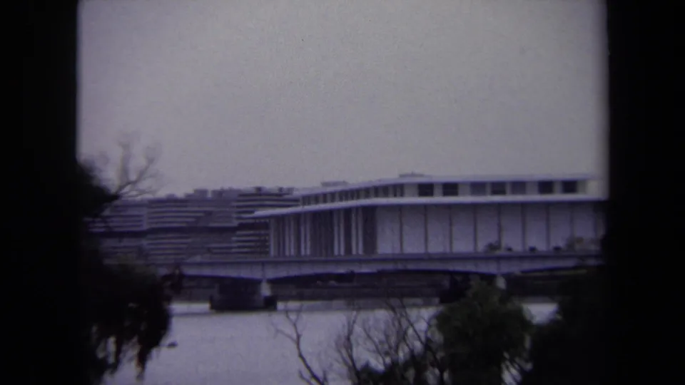 Zoomed 1970 Washington, DC view of a low bridge or overpass beside a modern riverfront building, partly obscured by trees.