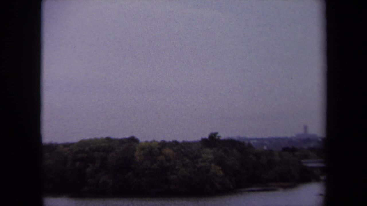 Tree-lined shoreline and broad river under a gray sky, framed by dark 8mm borders.
