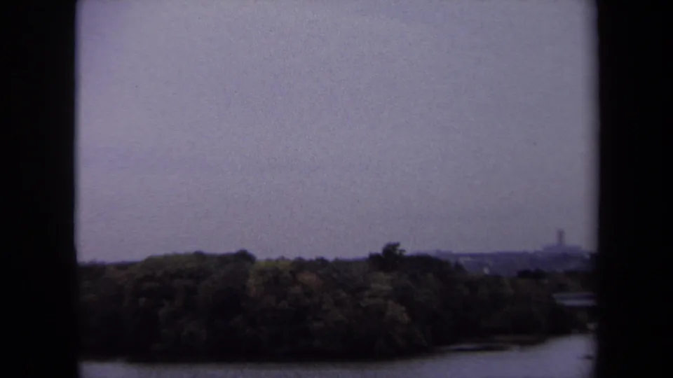 1970 Washington, DC area home-movie frame showing a wooded shoreline across a river beneath an overcast sky.