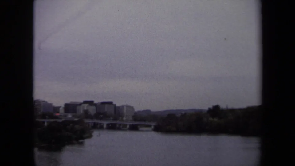 1970 Washington, DC area frame with low city buildings, a bridge, and river seen from a distant shoreline viewpoint.