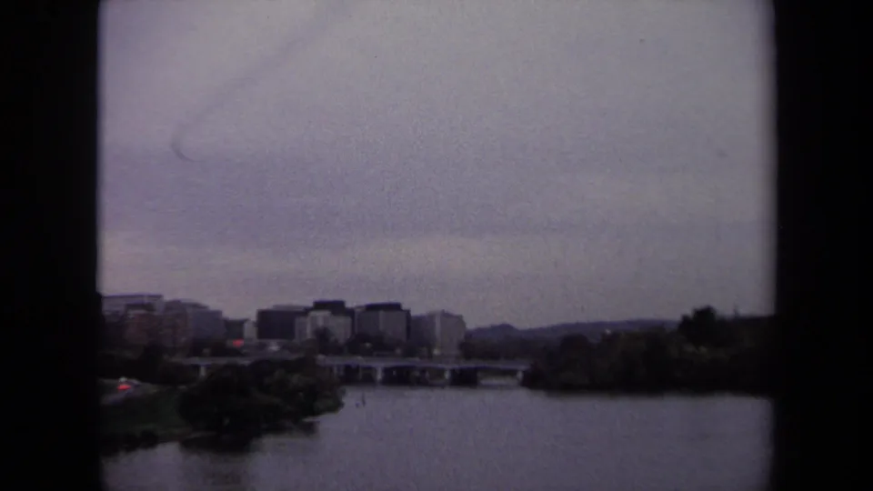 A Washington, DC area river view shows distant buildings and bridge with a faint aircraft trail across the gray 1970 sky.