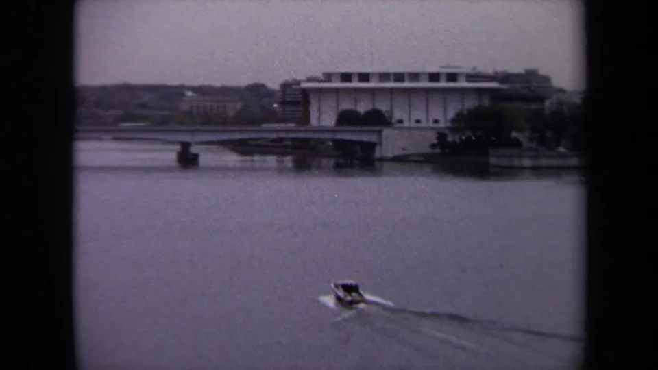 Washington DC waterfront view with a small boat below a bridge and large pale building, captured on grainy 8mm film.