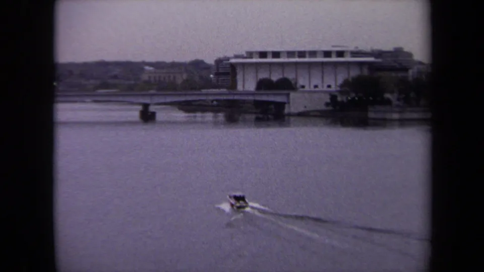 1970 Washington DC frame showing a small motorboat on a broad waterway with distant bridge and waterfront building.