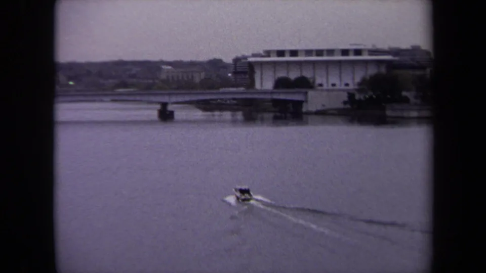 1970 8mm Washington, DC view of a small boat moving across water near a broad waterfront building and distant shore.