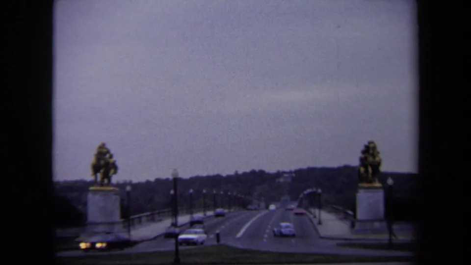 1970 Washington DC traffic moving along a broad bridge, framed by equestrian statues, lamp posts, and a distant tree-lined horizon.