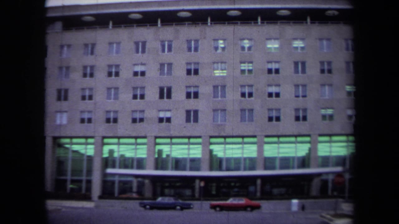 1970 Washington, DC view of the Department of State building facade with rows of windows, green-lit ground floor, and classic cars at the curb.