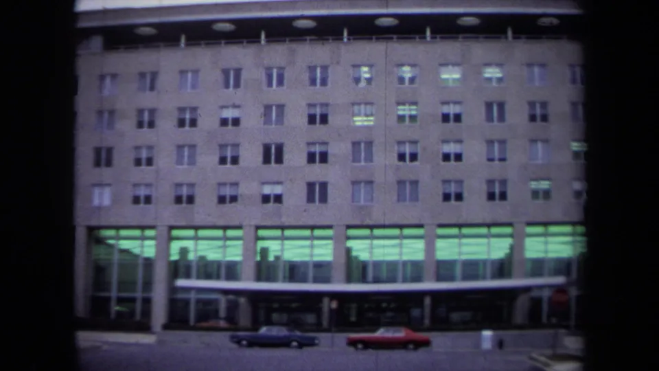 1970 Washington, DC view of the Department of State building facade with rows of windows, green-lit ground floor, and classic cars at the curb.
