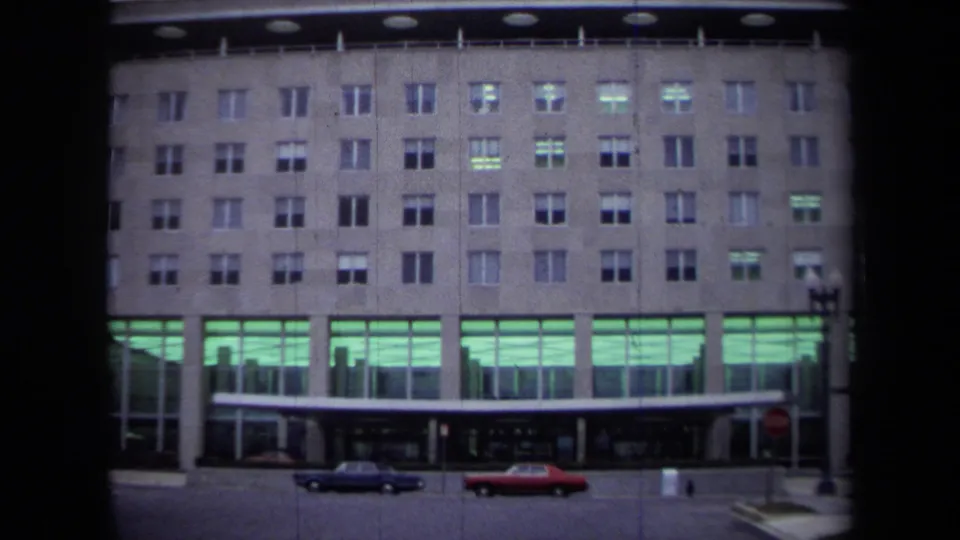 1970 Washington, DC exterior of the Department of State building, framed from across the street with a blue sedan and red car below the facade.