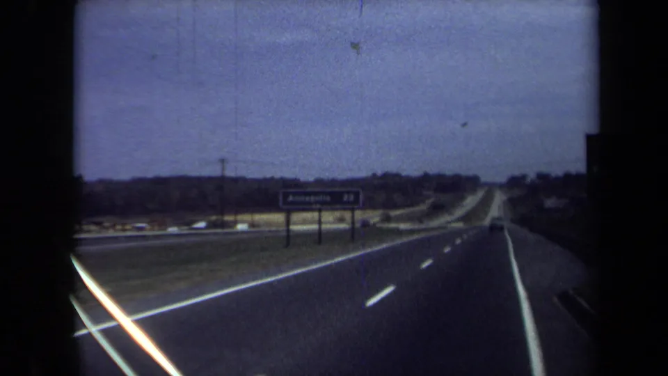 Passenger-view 1970 8mm footage looking down a highway toward an Annapolis direction sign under a muted sky.