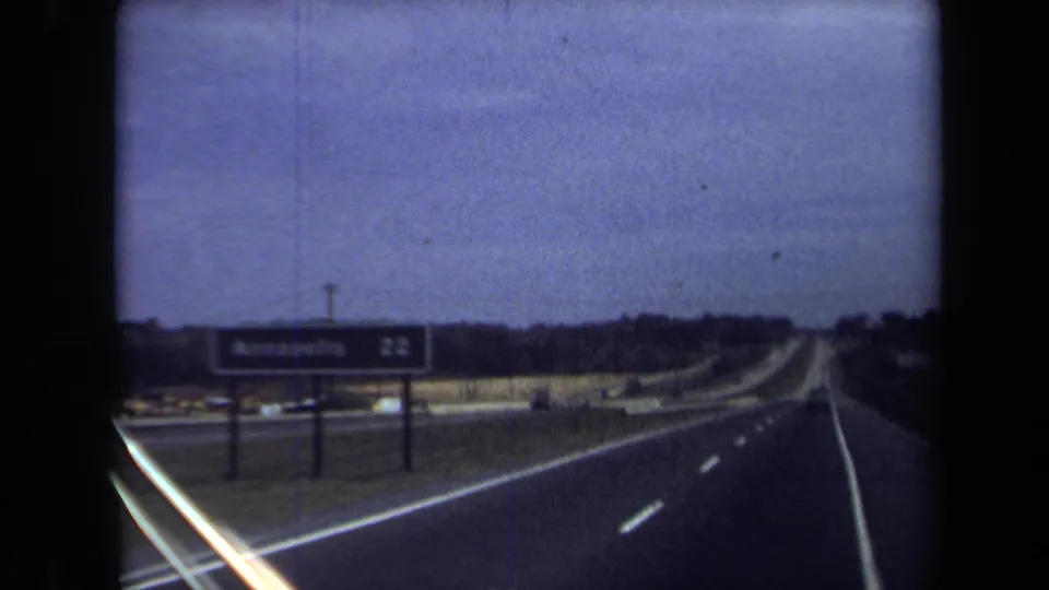Grainy 8mm passenger-view highway scene with road lanes, roadside signage, and low horizon after the Washington, DC exterior shot.