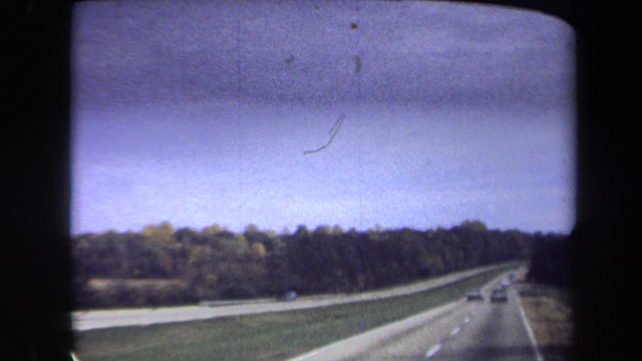 1970 Washington, DC 8mm frame showing a forward-moving view along a divided highway with trees, distant cars, and a dark mark in the sky area.