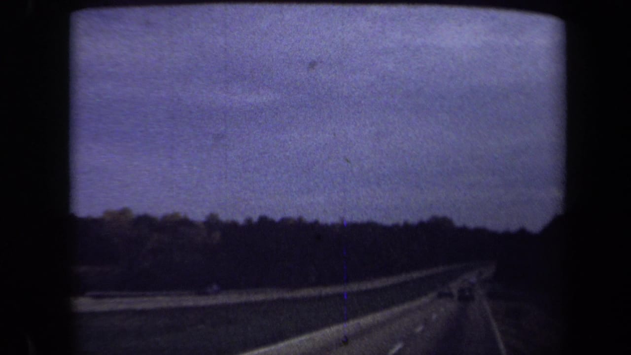 Dim highway view with tree line, roadway curve, distant vehicles, and darkened film exposure.