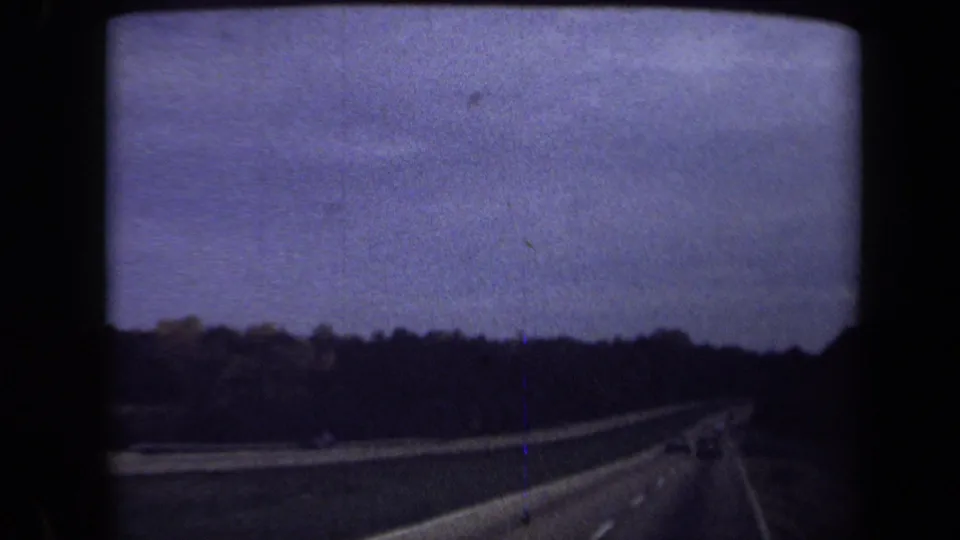1970 Washington, DC 8mm frame with a darker forward highway view, tree-lined road edges, distant cars, and visible film grain.