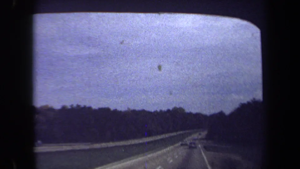 1970 Washington, DC 8mm frame showing a moving-vehicle view on a divided highway bordered by trees under a pale overcast sky.