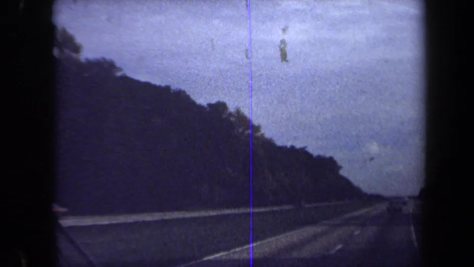 1970 Washington, DC 8mm frame showing a tree-lined highway edge, roadway lanes, distant traffic, and a vertical film scratch.