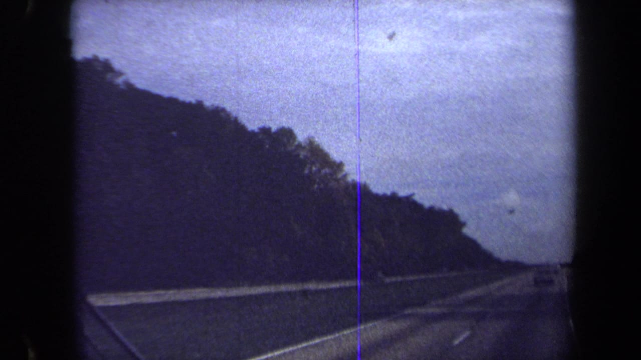 Forward view along a highway with trees on the left, guardrail, lane markings, and open sky.