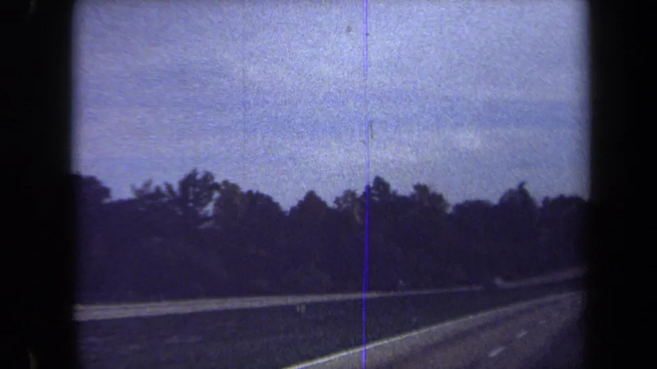 Later frame of the 1970 Washington, DC metadata highway drive, showing open lanes, roadside trees, guardrail, and pronounced 8mm film artifacts.