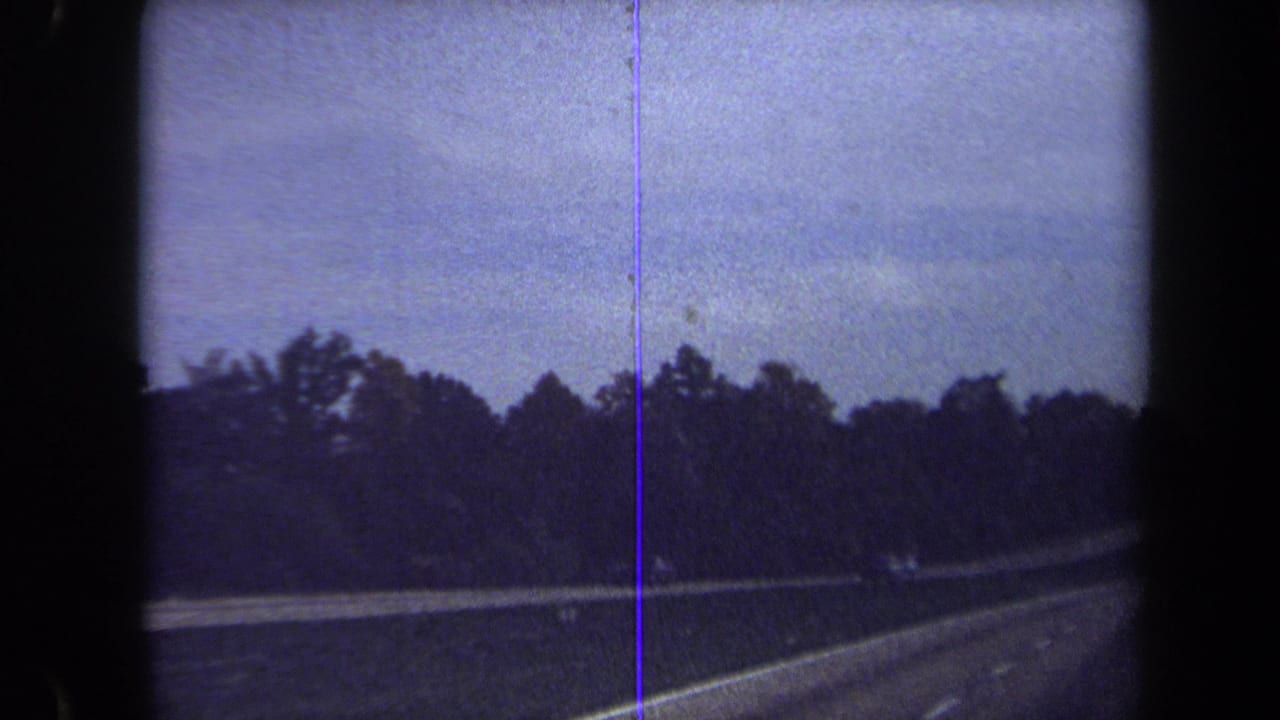 1970 Washington, DC area home-movie frame showing a curving road bordered by trees, guardrail, and a blue-tinted 8mm film edge.