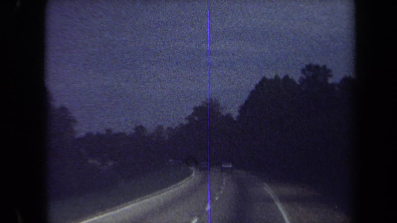 Blue-tinted 8mm frame from the Washington, DC area showing a wooded roadway curve, guardrail, and possible distant car ahead.