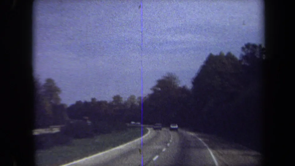 1970 Washington, DC area road view with several cars ahead, tree cover on both sides, lane stripes, and visible 8mm grain.