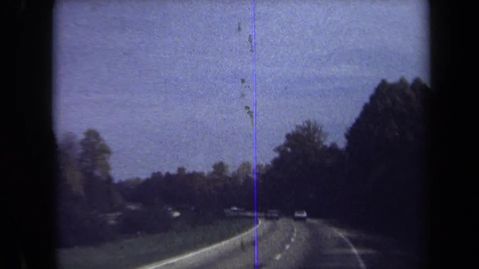 Washington, DC area home-movie frame showing cars traveling on a curving wooded road with lane markings and prominent film scratches.
