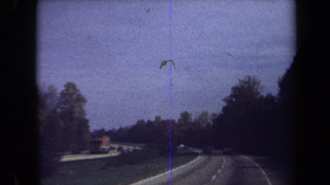 A 1970 home-movie frame shows a wooded Washington, DC-area highway curve with a truck and several cars ahead.